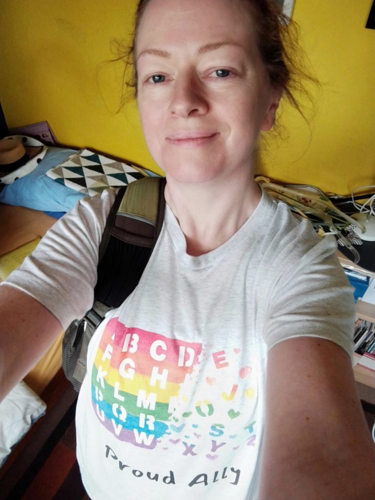 A selfie of awhite middle-aged female presenting person wearing a Spellers and Allies t-shirt and backpack against a yellow wall